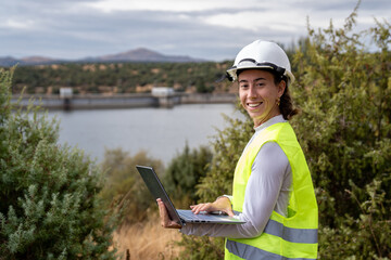 Female engineer wearing hard hat and safety vest, working on laptop near a dam, performing inspection and maintenance