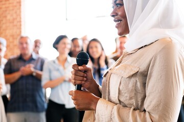 Woman in hijab holding a microphone in public with microphone, surrounded by diverse crowd. Joyful...