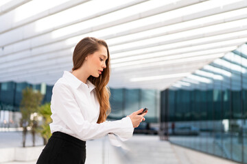 Young businesswoman with long brown hair wearing white shirt and black pants using smartphone while standing near modern office. She is focused on browsing in city during daytime