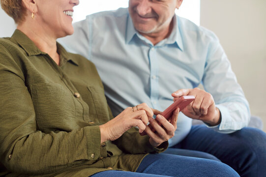 Happy elderly couple enjoying leisure time together at home using a phone for online entertainment and internet browsing. Their joyful communication reflects closeness and mutual happiness.