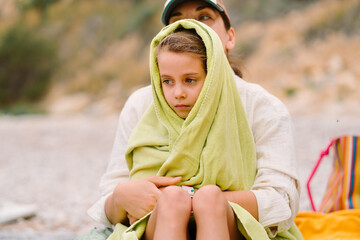 Mother embracing daughter wrapped in towel sitting on beach. The girl child looks calm and cozy while the mother provides comfort with serene beach background