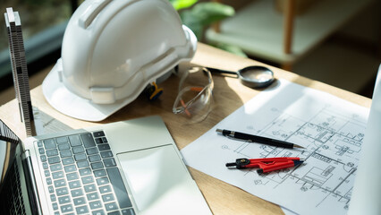 A white hard hat sits on a desk next to a laptop and a pair of safety glasses. A pair of red and black construction tools are also on the desk