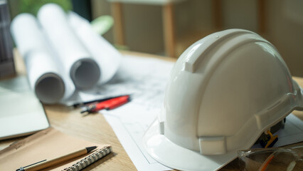 A white hard hat sits on a table with a stack of blueprints. The blueprints are for a construction project, and the hard hat is a safety measure for the workers
