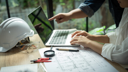 A man and a woman are working on a laptop together. The man is pointing at something on the screen. There are several construction tools on the table, including a compass and a pair of scissors
