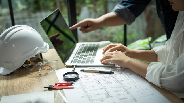 A man and a woman are working on a laptop together. The man is pointing at something on the screen. There are several construction tools on the table, including a compass and a pair of scissors - Powered by Adobe
