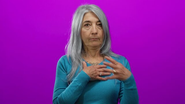 Senior woman with grey hair wearing blue sweater expresses surprise against isolated pink background with hand on chest conveying emotion.