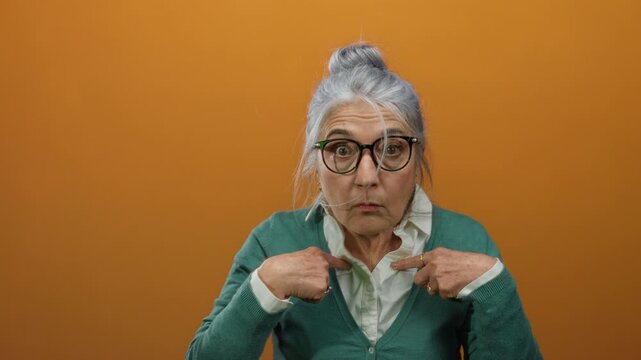Woman with grey hair and glasses pointing at herself, wearing a green cardigan against an orange background, expressing surprise and curiosity.