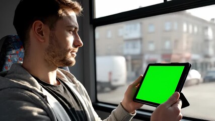 A man on a bus holding a tablet with a green screen, looking out the window. - Powered by Adobe