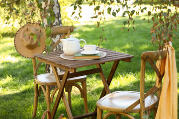 Comfortable wooden armchairs with book, cup and teapot on coffee table in park, outdoors