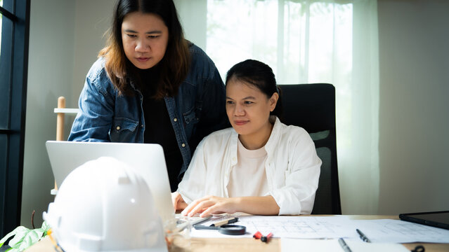 Two women are sitting at a desk looking at a laptop. One of them is wearing a hard hat