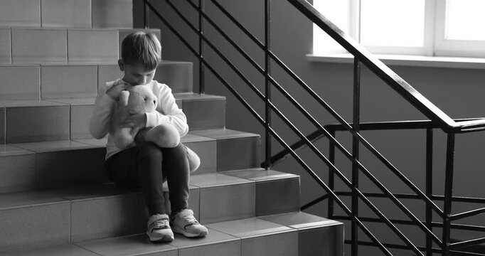 Upset little boy with toy bear on stairs indoors, black and white effect