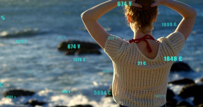 Gazing woman in white crocheted top gathering hair on rocky shore, with floating currency symbols - Powered by Adobe