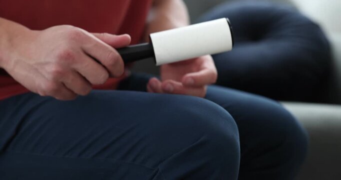 Man cleaning pants with lint roller on sofa at home, closeup
