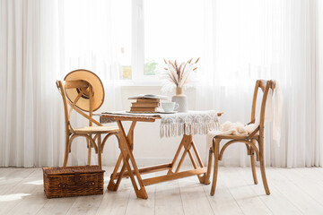 Interior of room with wooden chairs, wicker basket, cup of tea and books on table near window