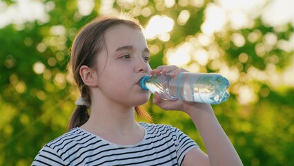Child girl drinks refreshing water from bottle, walk, park, nature. Beautiful girl drinks clean mineral water outdoors, summer heat in park. Exercise outdoors, quench your thirst with cool water.