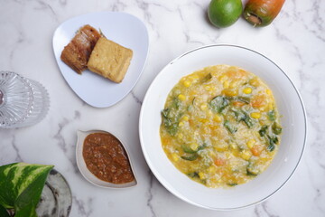 A traditional Indonesian meal featuring Bubur Manado or porridge with vegetables and salted fish on a classic plate, accompanied by fried tofu and a glass of water. A top view of traditional Bubur Man