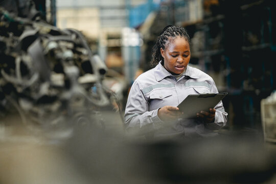 Female technician check used car damaged engine block at scrapyard warehouse recycle area part. African American engineer inspecting rust oily auto motor old spare part in junkyard for reuse service
