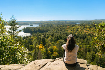 Woman sitting on rock overlooking Algonquin Park forest and lakes under clear blue sky.