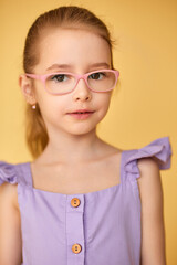Smart girl in pink glasses poses against yellow background. 
Child in lavender dress shows confidence and gentle charm.
Bright portrait radiates intelligence, style, and innocence.