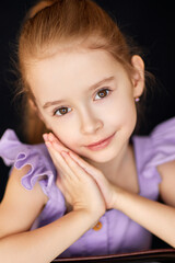 Adorable girl in lavender dress poses with gentle smile. 
Closeup child portrait shows innocence and natural beauty.
Soft light highlights graceful expression and bright eyes.