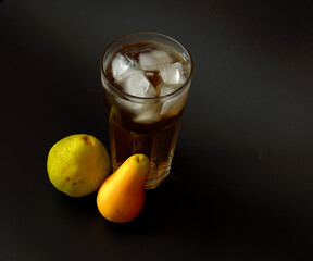 Pear juice in a tall faceted glass with ice on a black background, ripe fruits of different varieties nearby.