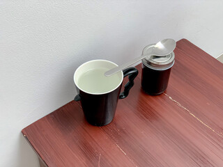 A black mug with liquid and a jar with a spoon on a wooden table against a white wall.