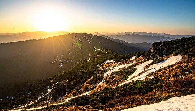 Majestic mountain range at sunset with golden sun rays illuminating sparse snow patches and evergreen trees under a clear sky in a vast wilderness