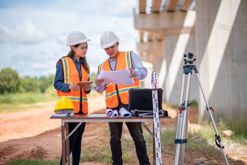 Engineers surveyor wearing helmets and reflective vests discuss construction blueprints work site surveying tripod and measurement equipment infrastructure or bridge construction industry.