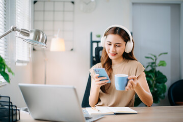 Asian woman working from home with laptop and headphones, smiling while using smartphone. and...