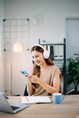 Asian woman working from home with laptop and headphones, smiling while using smartphone. and...
