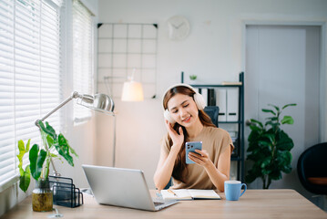 Asian woman working from home with laptop and headphones, smiling while using smartphone. and...