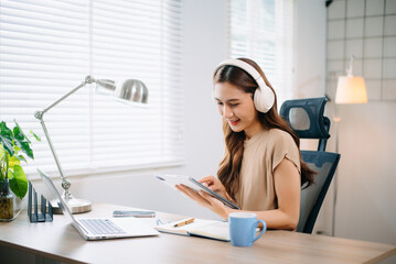 Young asian woman wearing headphones working with laptop and using smartphone while sitting at her...