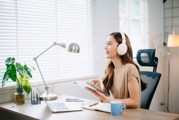 Young asian woman wearing headphones working with laptop and using smartphone while sitting at her...