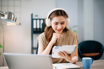 Young asian woman wearing headphones working with laptop and using smartphone while sitting at her...