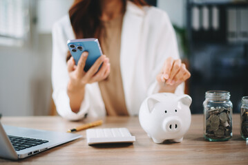 Asian woman smiling at camera holding piggy bank with coins and calculator on desk. Ideal for...