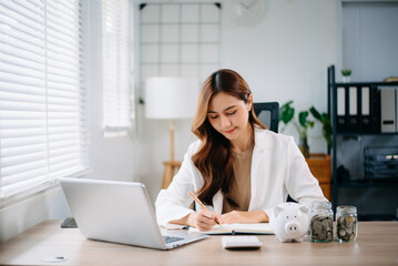 Asian woman smiling while managing finances at desk with laptop, clipboard, and coins.