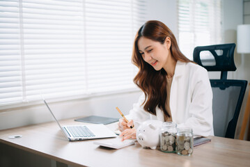 Asian woman smiling at camera holding piggy bank with coins and calculator on desk. Ideal for...