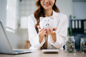 Asian woman smiling at camera holding piggy bank with coins and calculator on desk. Ideal for...