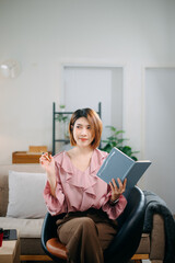 Young woman sits comfortably on a sofa, reading a notebook while holding a coffee cup.