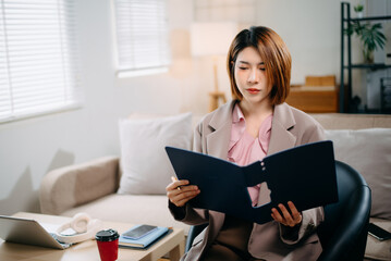 Asian woman holding paper and notebook on lap looking at laptop while sit on sofa