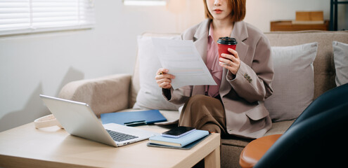Asian woman holding paper and notebook on lap looking at laptop while sit on sofa at office