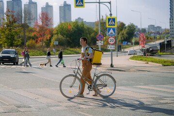 Bike courier crosses a busy city crosswalk on a bicycle with a delivery backpack, providing fast, professional urban food delivery amid pedestrian and traffic activity