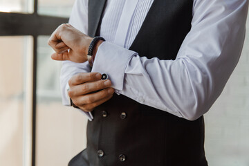 A young Caucasian man adjusts his cufflinks while wearing a formal black vest and white shirt. He...