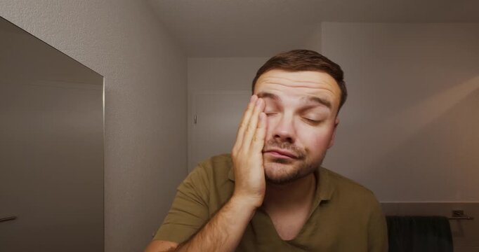 A sleepy man stands in front of the bathroom mirror, looking exhausted and unmotivated as he rubs his face and struggles to wake up &mdash; a realistic morning-after or hangover moment.