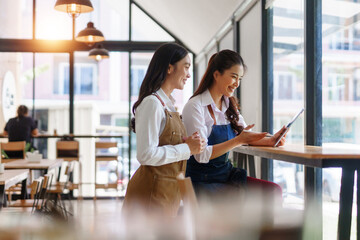 Young Asian women baristas discussing business, smiling and using a digital tablet inside a modern...
