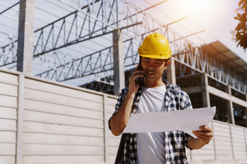 Asian man in hard hat reviewing construction blueprint while talking on smartphone at an active...