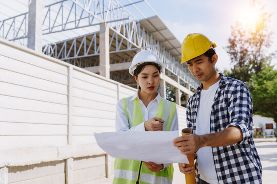 Two Asian engineers wearing hard hats reviewing a construction blueprint at a building site, planning their project