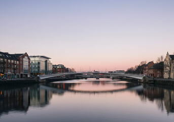 Obraz premium Peaceful urban cityscape with a prominent bridge arching over a placid river, reflecting the gentle colors of dusk