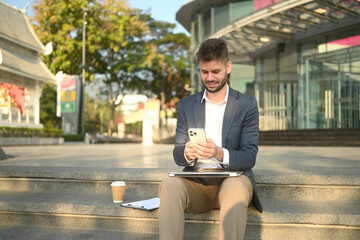 Caucasian man in business attire sitting on outdoor steps using smartphone, working remotely with laptop and coffee