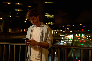 Young man connecting with phone on city bridge at night